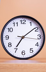 Minimalist black-framed analog clock displaying the time 7:08, set against neutral beige background and resting on wooden surface.