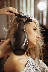 Makeup artist hairdresser combing hair of female model with comb and hair dryer. Close-up vertical view