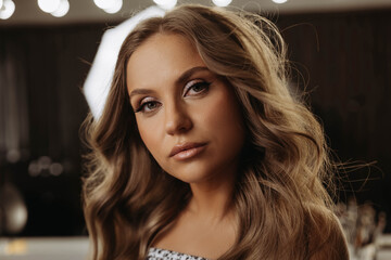 Portrait of attractive young woman with wavy hair. Behind woman there is mirror and lights of beauty salon. Model looks expressively at camera. Close-up