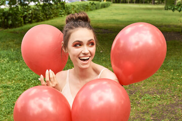 Young caucasian adult female smiling surrounded by red balloons in green outdoor park setting.