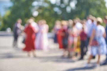 Blurred crowd of people socializing outdoors in sunlit urban setting with lush green trees.