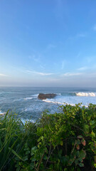 a wide view of a large coral rock in the middle of the sea exposed to many sea waves and a blue sky with thin clouds