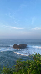 a wide view of a large coral rock in the middle of the sea exposed to many sea waves and a blue sky with thin clouds