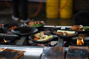 A close-up shot of several plates of Indonesian Ayam Penyet (smashed fried chicken with sambal) on a rustic street food stove. The flames below are heating the food for serving. Authentic outdoor cook