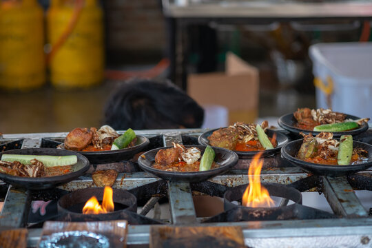 A close-up shot of several plates of Indonesian Ayam Penyet (smashed fried chicken with sambal) on a rustic street food stove. The flames below are heating the food for serving. Authentic outdoor cook