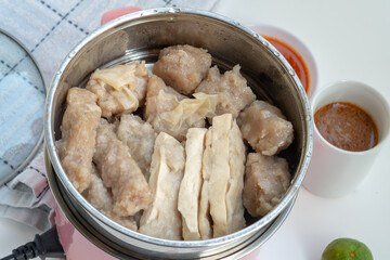 Various steamed Indonesian siomay and tofu pieces are shown in a metal pot, with cups of peanut sauce and chili sauce in the blurred background.