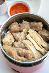 Various steamed Indonesian siomay and tofu pieces are shown in a metal pot, with cups of peanut sauce and chili sauce in the blurred background.