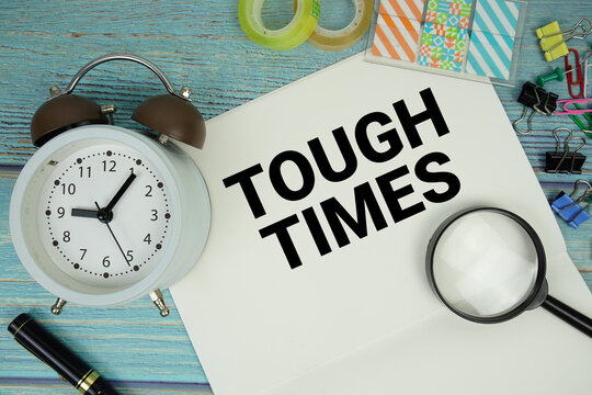 Tough Times text on a paper surrounded by office supplies, a magnifying glass, and a clock on a blue wooden surface