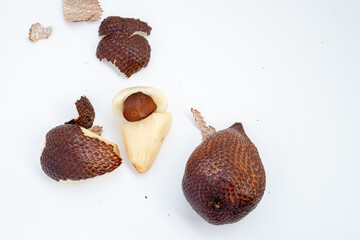 A few spiky brown salak fruit, one peeled to reveal the white flesh and a dark brown seed, are arranged on a clean white surface with some loose pieces of peel.