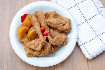 A plate of Indonesian braised chicken and potatoes in a brown sauce, resembling a chicken curry, garnished with red chili peppers, sits on a wooden table.