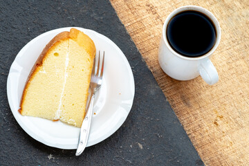  A single slice of simple cake with a fork on a white plate, positioned on a diagonally split black and wooden background.