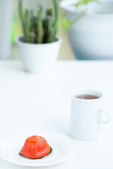 A small, red, tortoise-shaped Chinese sweet dumpling, known as ang ku kueh, sits on a white plate next to a white mug of tea. The background is a clean, bright, out-of-focus scene with a potted cactus