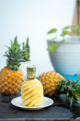 A bright, high-key photo of a peeled pineapple (Ananas comosus) with a spiral cut. The fresh, juicy fruit sits on a white plate, with whole pineapples and green plants creating a soft bokeh background
