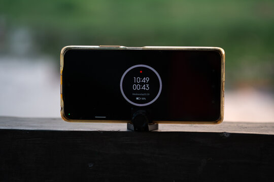 A gold smartphone rests horizontally on a small black stand, sitting on a dark wooden railing. The screen shows a digital clock and timer at 10:49 with 55% battery, set against a blurry green backgrou