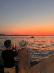 rear view of man and woman standing together taking photo watching sunset over Adriatic Sea in...