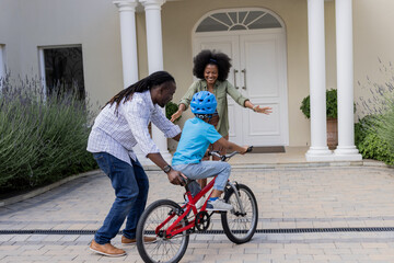 Naklejka premium African American parents teaching child to ride bicycle outside home, all smiling