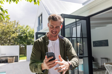 Smiling man using smartphone outside modern home, enjoying sunny day
