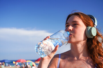 Young woman in headphones drinks from water bottle at sunny beach. Background with umbrellas and blurred shoreline.