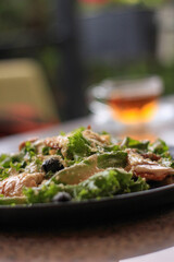 Close-up of chicken Caesar salad with greens, grilled chicken, olives, and a blurred iced tea.