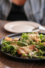 Caesar salad with greens, croutons, cheese on black plate, with a white plate and person in background.