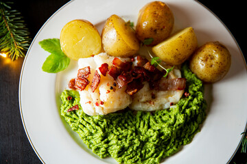 Traditional Christmas lutefisk dinner with pea puree, potatoes and bacon, served on porcelain plate with candles and spruce branches.