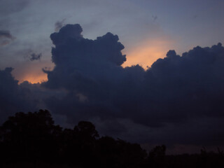 A dramatic, dark cloud formation silhouetted against the warm glow of a sunset sky above a treeline.