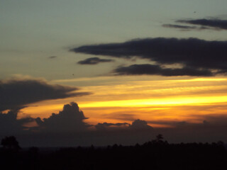 A dramatic sunset sky with vibrant yellow and orange light glowing behind dark silhouetted clouds over a landscape.