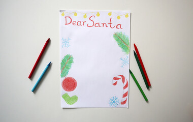 Children's drawing of Dear Santa letter with drawings of fir branches, Christmas ornaments, snowflakes, candy cane on white surface. On white table, surrounded by colored pencils. Top view. Template.
