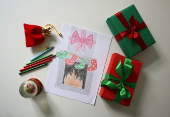 Child's drawing of a fireplace with Christmas stockings, placed on a white surface. Surrounded by festive gifts, snow globes, pencils on white table. Gifts in red and green colors with ribbon bows.