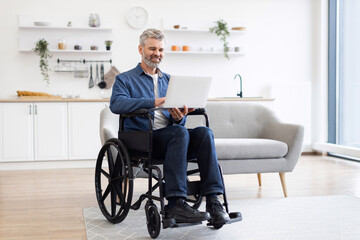 Mature Caucasian man with gray hair sitting in wheelchair working on laptop indoors. Bright and modern home setting emphasizing productivity, technology use, and independence.