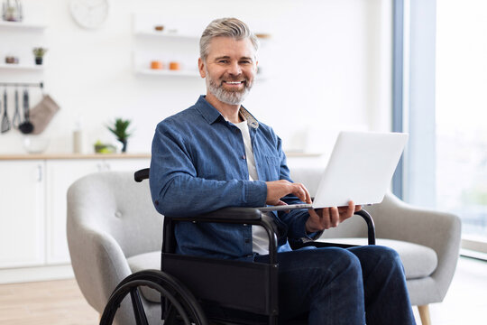 Mature Caucasian man sitting in wheelchair using laptop in bright spacious interior. Scene shows productivity, determination, and inclusivity in modern environment.