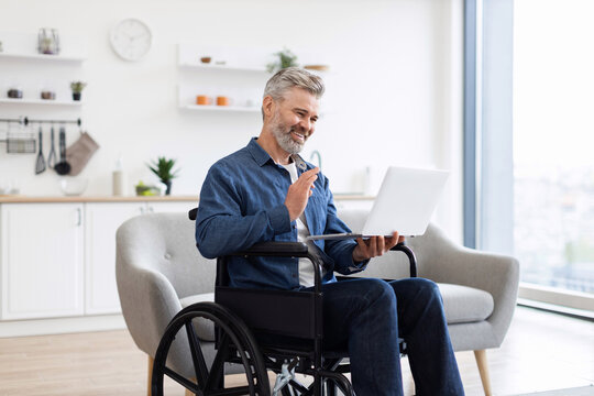 Mature Caucasian man using laptop in wheelchair having video conversation in bright home. Laptop in hands, engaged in communication. Wheelchair in modern interior showcasing accessibility