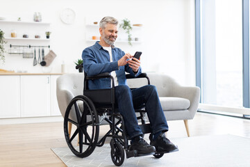 Mature Caucasian man sitting in wheelchair in home interior, holding smartphone, browsing.