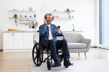 Mature Caucasian man in wheelchair using smartphone, listening to music, and smiling in bright living room. Wearing casual outfit with headphones, enjoying moment of relaxation and happiness indoors.