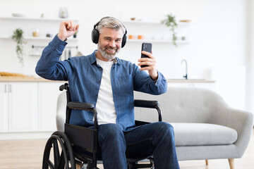 Mature Caucasian man in wheelchair listening to music with headphones using smartphone. Expresses joy, independence, technology. Captures indoors moment of lifestyle, entertainment