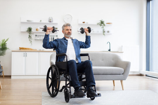 Mature Caucasian man in wheelchair lifting dumbbells during fitness activity at home, showcasing positivity, strength, and resilience. Bright and modern environment with natural lighting
