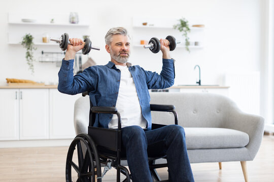 Mature Caucasian man sitting in wheelchair performing exercise with dumbbells in bright home interior, promoting fitness and determination.