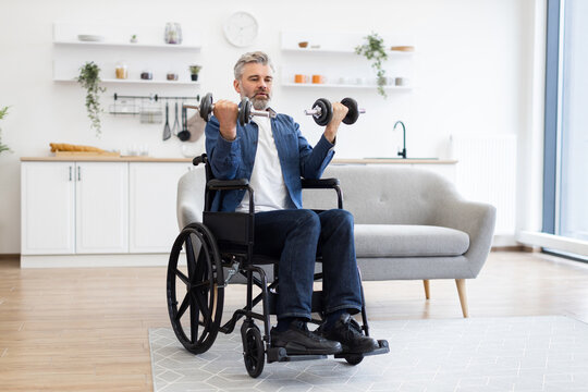 Mature Caucasian man seated in wheelchair, performing a workout with dumbbells indoors. Middle-aged adult engaged in strength training, promoting physical fitness and healthy lifestyle
