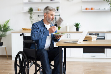 Middle-aged man sitting in wheelchair at table, eating salad and using laptop computer in contemporary kitchen space. Concept of independence, healthy living, modern lifestyle, and remote work setup.