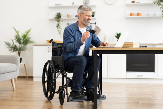 Mature man in wheelchair using laptop for online payment with bank card inside modern home setting. Concept of accessibility, financial independence, and modern technology for everyday transactions. - Powered by Adobe