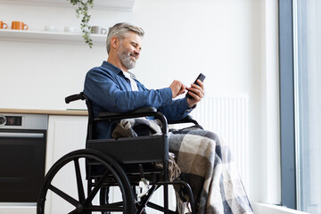 Mature man in wheelchair with plaid blanket using smartphone indoors in bright modern kitchen