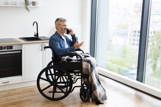 Elderly man sitting in wheelchair in cozy home using smartphone for communication. Bright environment with modern kitchen and large window providing natural light.