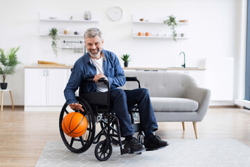 Gray-haired man wearing denim and white shirt in wheelchair practicing with basketball, indoors.