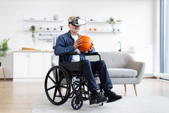 Senior man sitting in wheelchair indoors wears VR headset holding basketball, enjoying technology. Interior setting includes sofa, shelves. Emphasizes virtual reality, accessibility