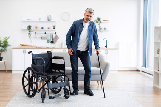 Middle-aged white man with disability using cane in home setting, wheelchair nearby. Depicts determination, recovery, mobility aids, progress within comfortable contemporary setting