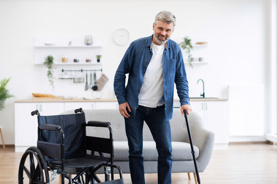 Middle-aged white man with disability using cane in home setting, wheelchair nearby. Depicts determination, recovery, mobility aids, progress within comfortable contemporary setting