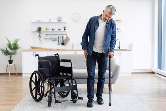 Mature disabled man learning to walk with assistance of cane, standing by wheelchair. Bright modern home interior, independence, recovery. Middle-aged male showing progress in rehabilitation at home.