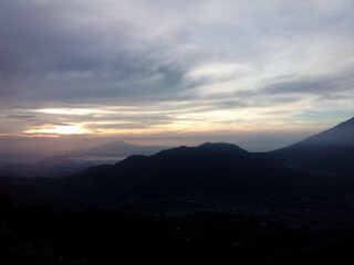 Obraz premium A dramatic sunrise over silhouetted mountain ranges under a cloudy, atmospheric sky.
