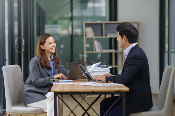 Two Asian businesspeople smiling and engaging in a strategic discussion while using a laptop in a modern office environment