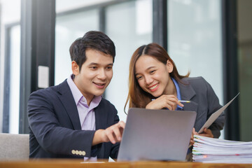 Two Asian business professionals smiling and collaborating while working together on a laptop in a modern office environment
